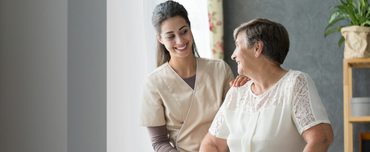 nurse standing next to elderly patient with her hand on her shoulder and both women are smiling