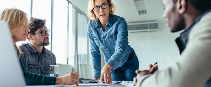 standing woman with glasses in office setting talking to co-workers that are sitting